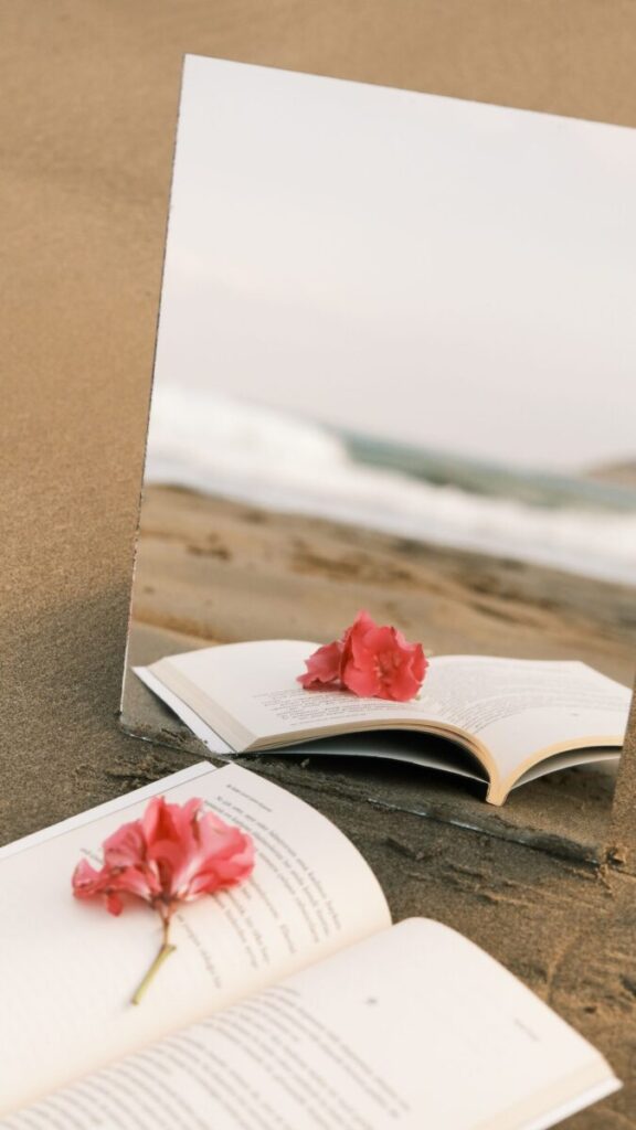 A creative setup featuring a mirror on the beach reflecting an open book and pink flowers.